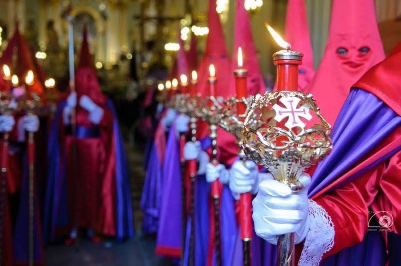 Fila de cofrades de «Los Azotes» con túnica y capirote rojos, capa morada y guantes blancos, portando los ciriales rematados con la cruz del escudo.